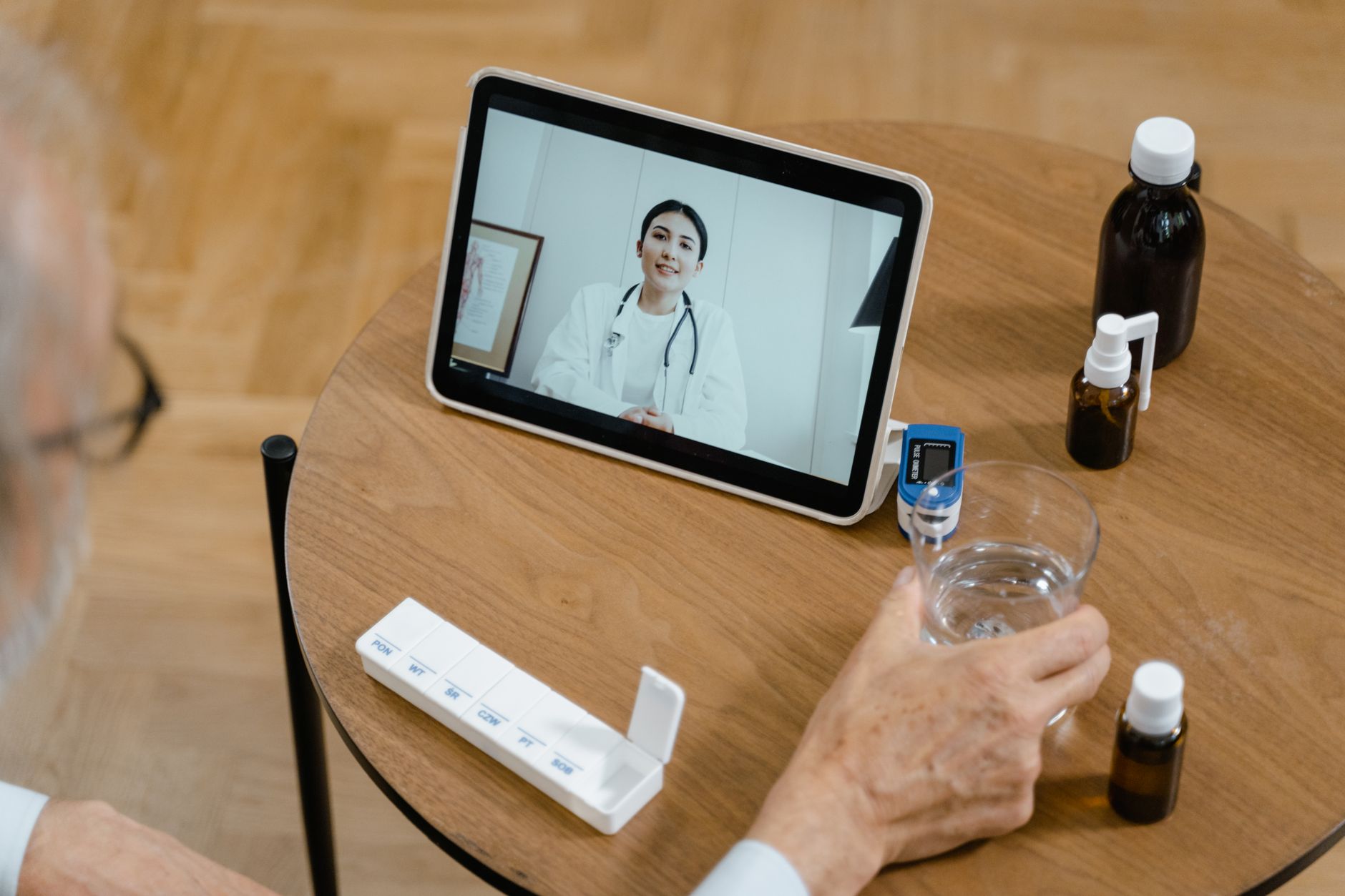 Persona frente a una tableta durante una videollamada con una profesional de salud, junto a medicamentos y un organizador semanal.