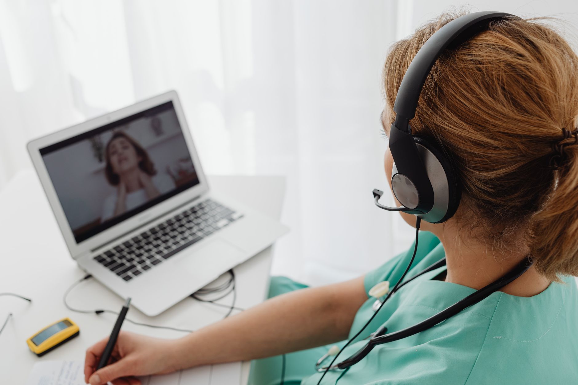 Una persona joven pensativa, revisando notas en un cuaderno después de una consulta médica virtual en Costa Rica, con una computadora y un teléfono cerca.
